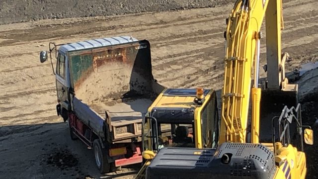 シャベルカーがトラックの荷台へ、車体を旋回させて、土を運ぶ写真
